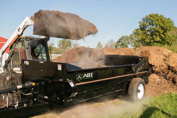 A 185ft³ PTO manure spreader being loaded by a skid steer bucket.